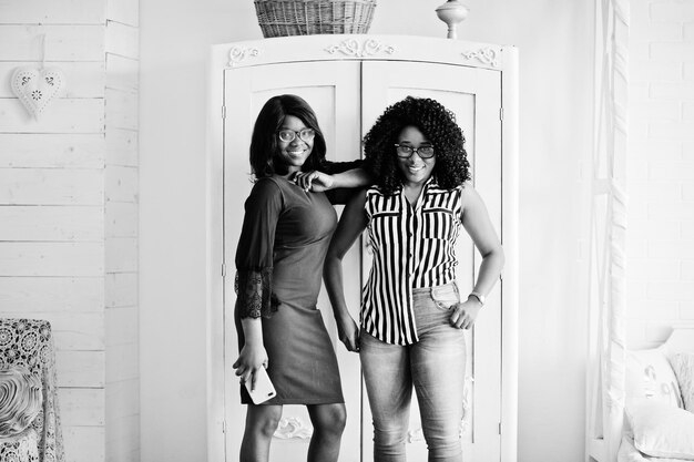 Two african woman friends wear on eyeglasses posed indoor white room against rustic wardrobe One of them hold mobile phone at hand