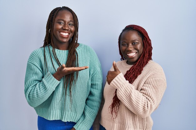 Two african woman standing over blue background showing palm hand and doing ok gesture with thumbs up, smiling happy and cheerful