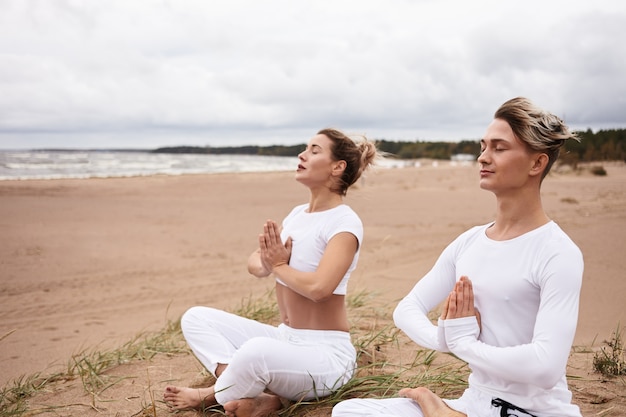 Free photo two atheltic european people man and woman in white sportswear closing eyes and holding hands in namste gesture, sitting in padmasana while meditating outdoors during yoga retreat by the ocean