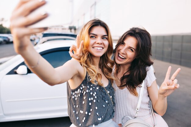 Two attractive fashionable girls having fun on parking. They making selfie-portrait and look happy.