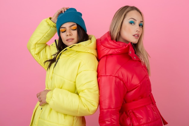 Two attractive girls posing on pink background in colorful winter down jacket of bright red and yellow color