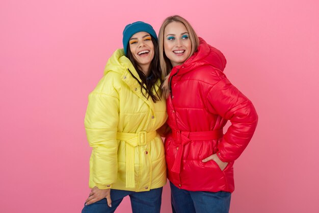 Two attractive girls posing on pink background in colorful winter down jacket of bright red and yellow color