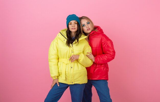 Two attractive stylish women posing on pink background in colorful winter down jacket of red and yellow color, warm clothes fashion trend