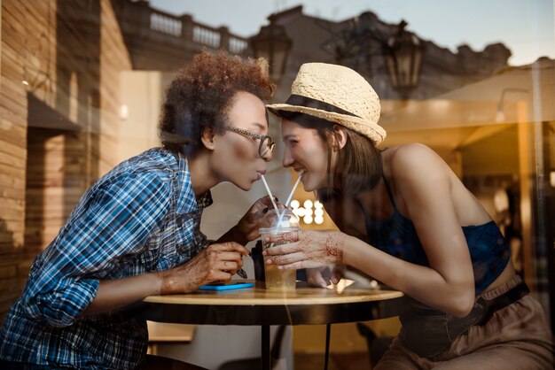 Two beautiful girls smiling, drinking from tubes, resting in cafe. Outside shot.