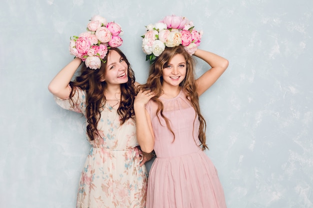 Free photo two beautiful girls stand in a studio, play silly and have circlets of flowers on their heads.