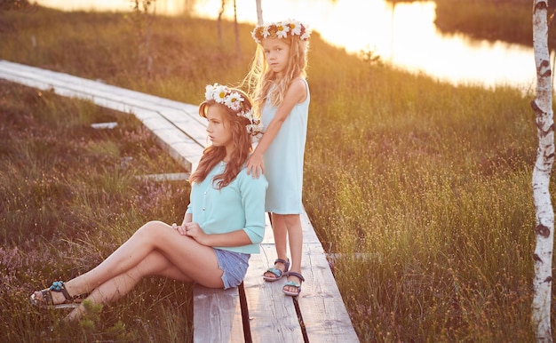 Free Photo two beautiful sisters standing against the background of a beautiful landscape, walk on the field near a pond at bright sunset.