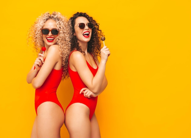 Two beautiful smiling hipster women in red swimwear bathing suitsTrendy models with curls hairstyle in studio Hot female posing near yellow wall in sunglassesHolding lollipop Isolated on white