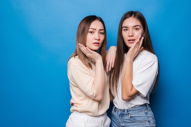 Two beautiful surprised screaming with their hands up, dressed in casual clothes on a blue wall. People emotions concept.