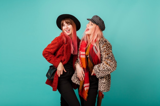 Two beautiful women in stylish faux fur coats and wool scarf posing on turquoise wall