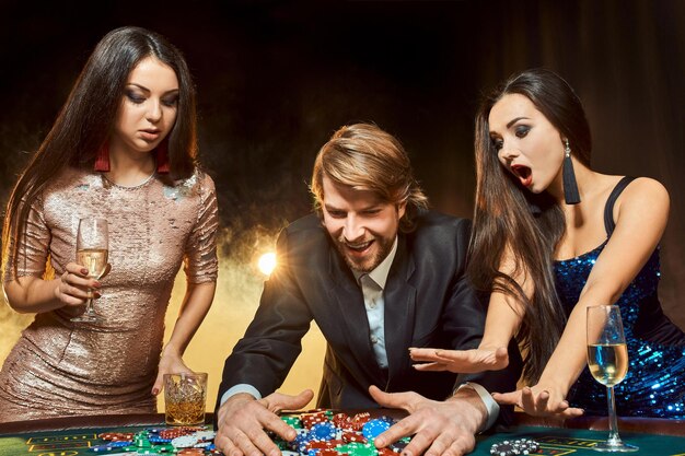 Two beautiful women and young man play on poker table in casino, focus on man and brunette. Passion, cards, chips, alcohol, dice, gambling, casino - it is entertainment. Dangerous fun card game for mo