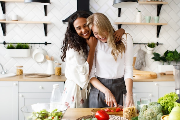 Free photo two beautiful young women are making a healthy breakfast and hugging near the table full of fresh vegetables on the white modern kitchen