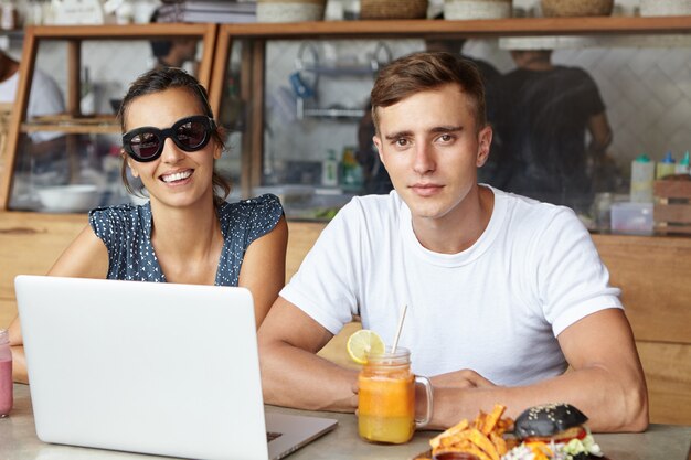 Two best friends using laptop computer during lunch, sitting in cozy cafe interior and looking with happy smiles. Students studying online on notebook pc