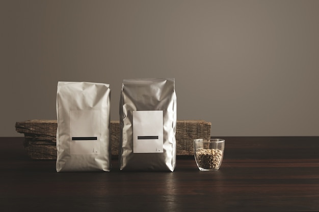 Two big hermetic packages with blank labels near transparent glass with raw sampled coffee beans