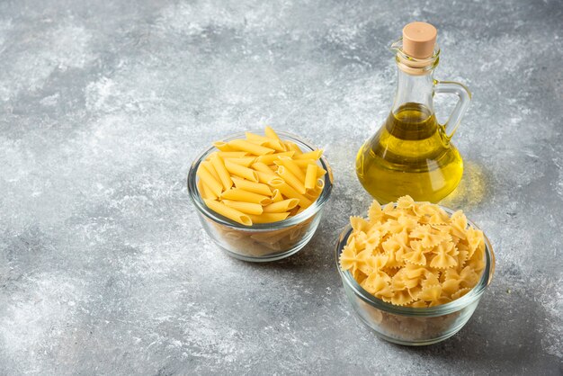 Two bowls of raw penne and farfalle pasta with bottle of olive oil on marble background. 