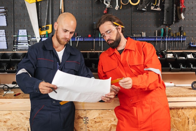 Free photo two builders in work clothes thoughtfully looking on sketch plan with tools on background in workshop