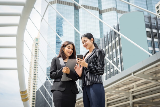 Two business woman standing using smartphone and discussing in front of the office. Business working concept.