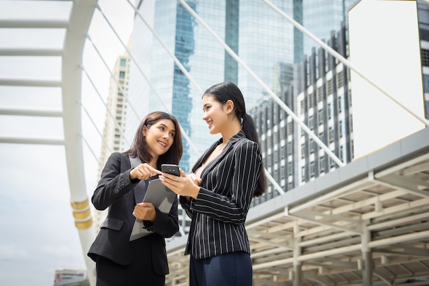 Two business woman standing using smartphone and discussing in front of the office. Business working concept.