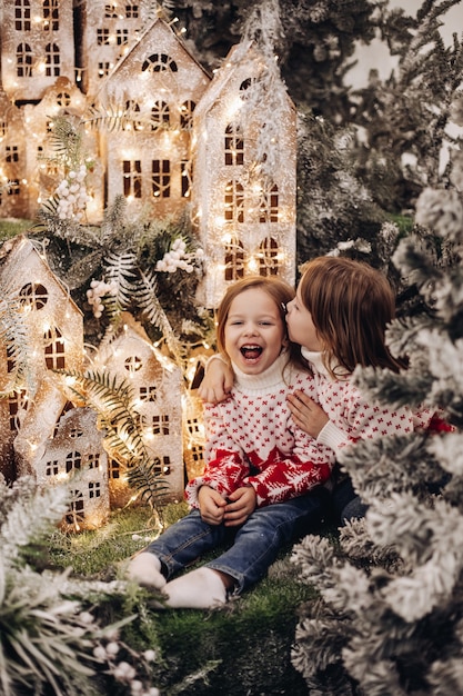 Free photo two caucasian sisters poses for the camera in a beautiful christmas decoration with a lot of trees undder a snow