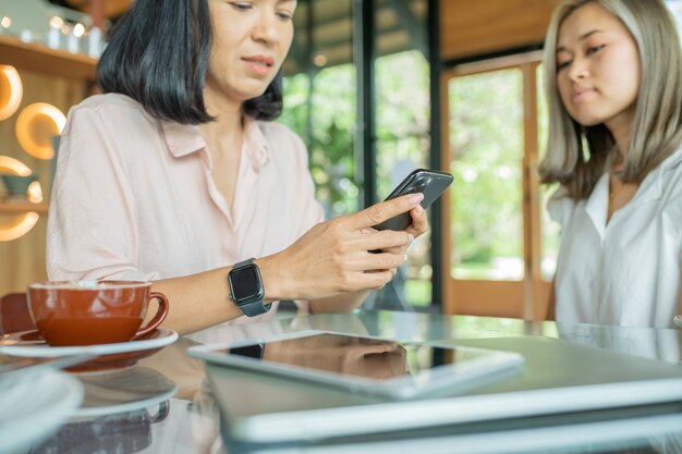 Two cheerful and beautiful girls are sitting together near the table and watching something on the phone. they look relaxed and happy. also girls are enjoying the time spending together.