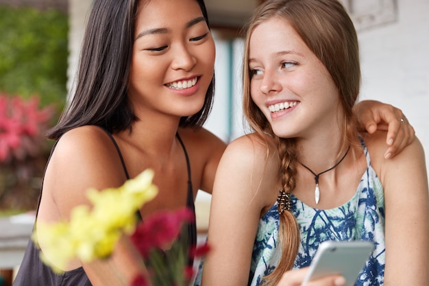 Two cheerful girlfriends of different nationalities, embrace, have fun together, rest in cafe, hold modern smart phone