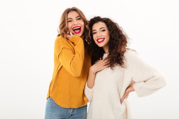 Two cheerful girls in sweaters posing together  over white wall