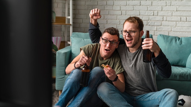 Free photo two cheerful male friends watching sports on tv and having beer