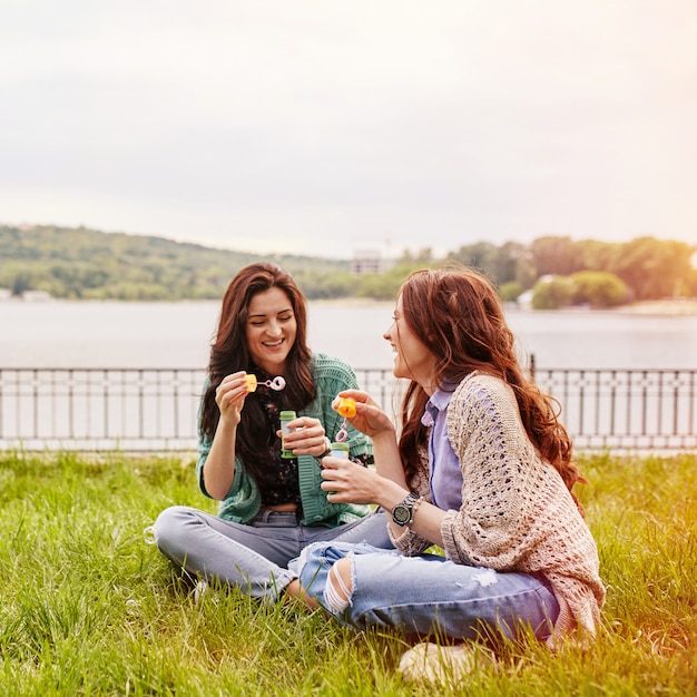 Two cheerful sisters sitting on the grass and blowing bubbles