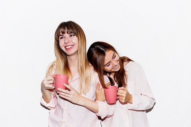 Two cheerful white  women in pink pajamas with cup of tea posing. Flash portrait.