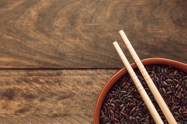 Two chopsticks over the black rice grain bowl on wooden background