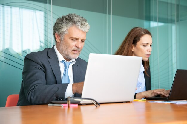 Two colleagues sitting together and using computers in office. Employees of different ages typing on laptop keyboards.