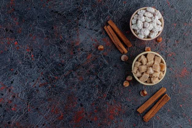 Two colorful bowls full of sweet white and brown candies with healthy nuts and cinnamon sticks . 