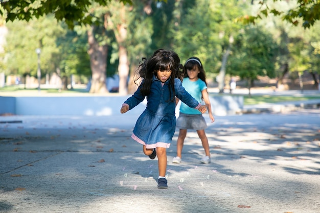 Two excited black haired little girls playing hopscotch in city park. Full length, copy space. Childhood concept