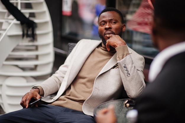 Two fashion black men sitting outdoor with mobile phone Fashionable portrait of african american male models Wear suit coat and hat