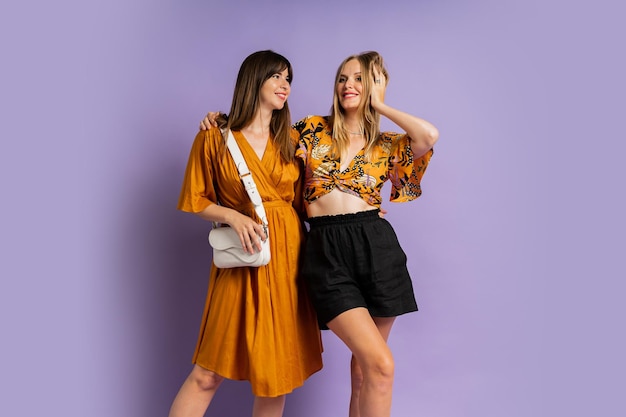 Two fashionable european women posing in studio over purple background Wearing stylish summer outfit