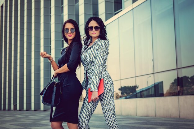 Free photo two fashionable women in stylish clothes and sunglasses posing in a middle of business urban district.