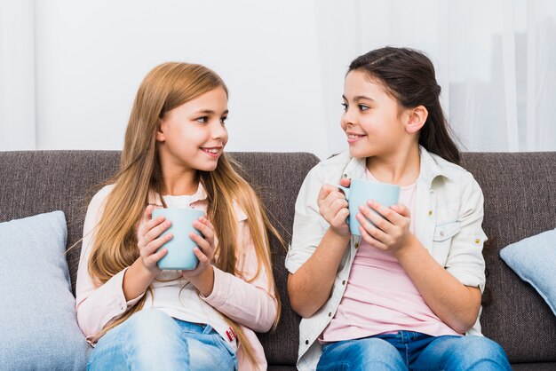 Two female friends sitting on sofa holding coffee mug in hand looking at each other