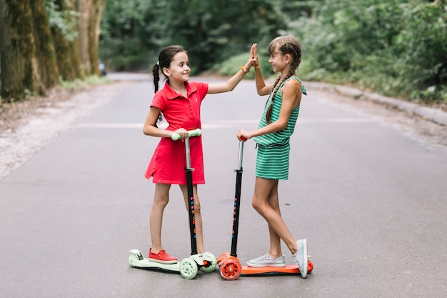 Free photo two female friends standing on scooter giving high five gesture on road