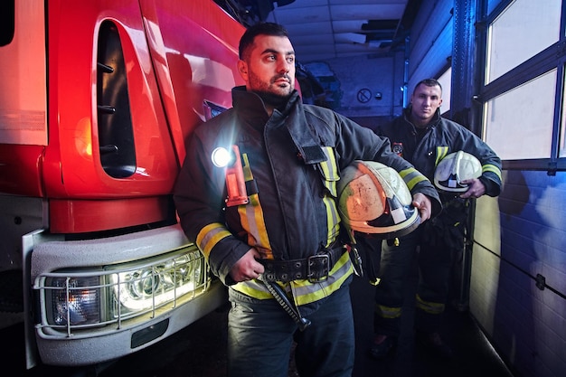 Two firemen wearing protective uniform standing next to a fire engine in a garage of a fire department.