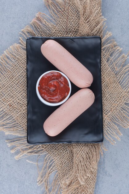 Two fresh boiled sausage with ketchup on black plate. 