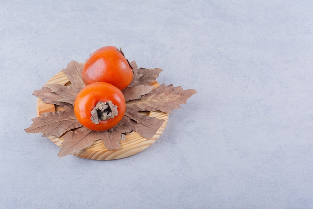 Free Photo two fresh persimmon fruits and dried leaves on wooden plate.