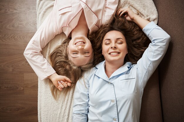 Two friends lying on sofa in cozy pyjamas with closed eyes, smiling and relaxing