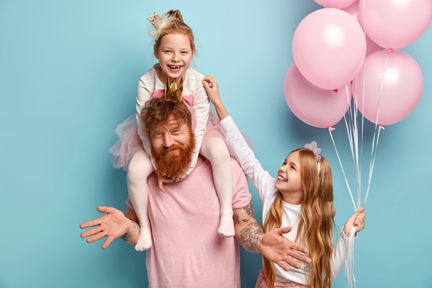 Free photo two funny naughty sisters play together, tickle each other being cared by dad. cute little girl holds bunch of air balloons. happy family of father and two daughters prepare for holiday, pose indoor