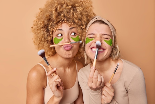 Free photo two funny women apply facial powder with cosmetic brushes undrgo beauty procedures use green hydrogel patches under eyes stand next to each other isolated over brown background skin care concept