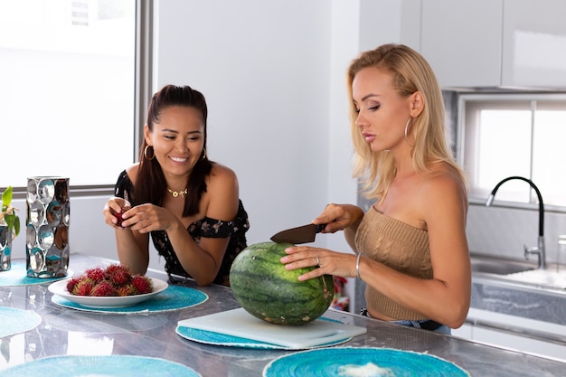 Free Photo two girl friends eating watermelon and rambutan tropical fruits in kitchen