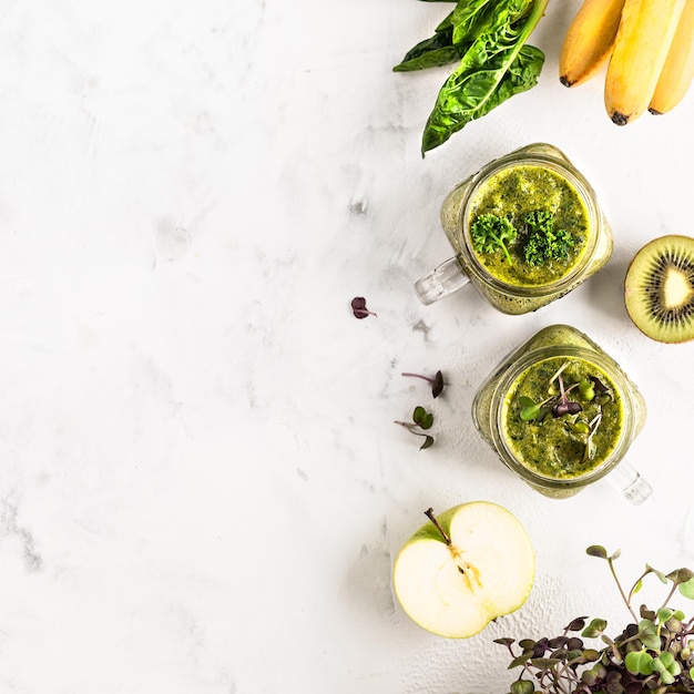 Two glass jars of homemade spinachbased green cocktail with banana apple and kiwi and wet greens on a white background top view