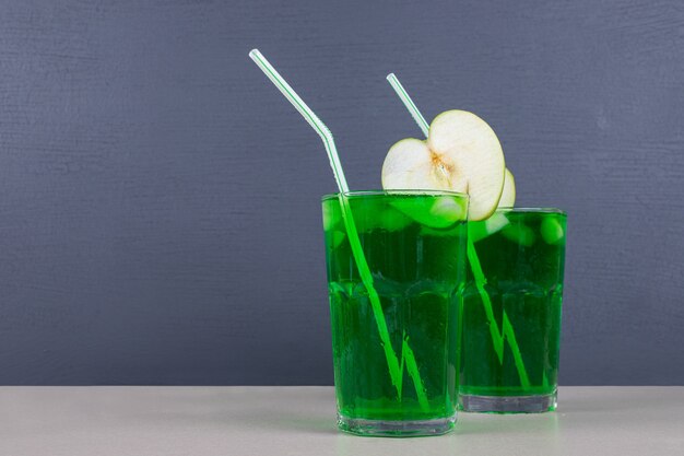 Two glasses of green juice with straws on blue table.