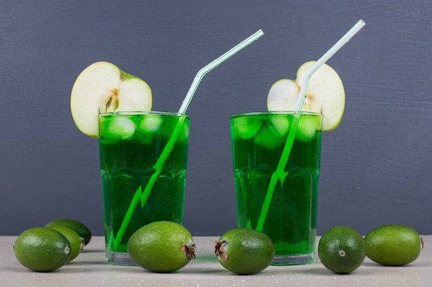 Two glasses of green juice with straws on blue wall.