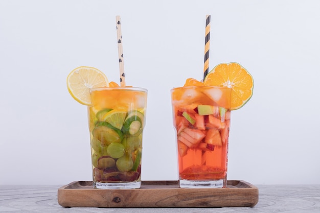 Two glasses of juice with fruit slices on white wall.