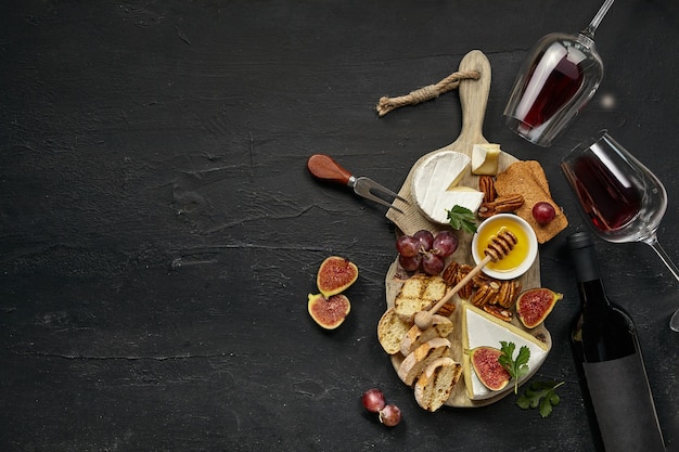 Free Photo two glasses of red wine and a tasty cheese plate with fruit, grape, nuts and toasted bread on a wooden kitchen plate on the black stone background