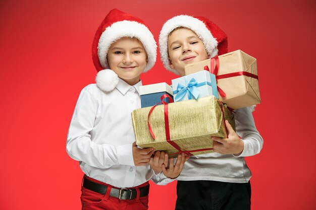 Two happy boys in santa claus hats with gift boxes on red